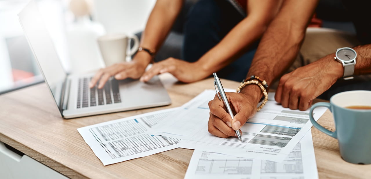 Hands working on a laptop and filling out financial documents spread across a desk with coffee nearby.